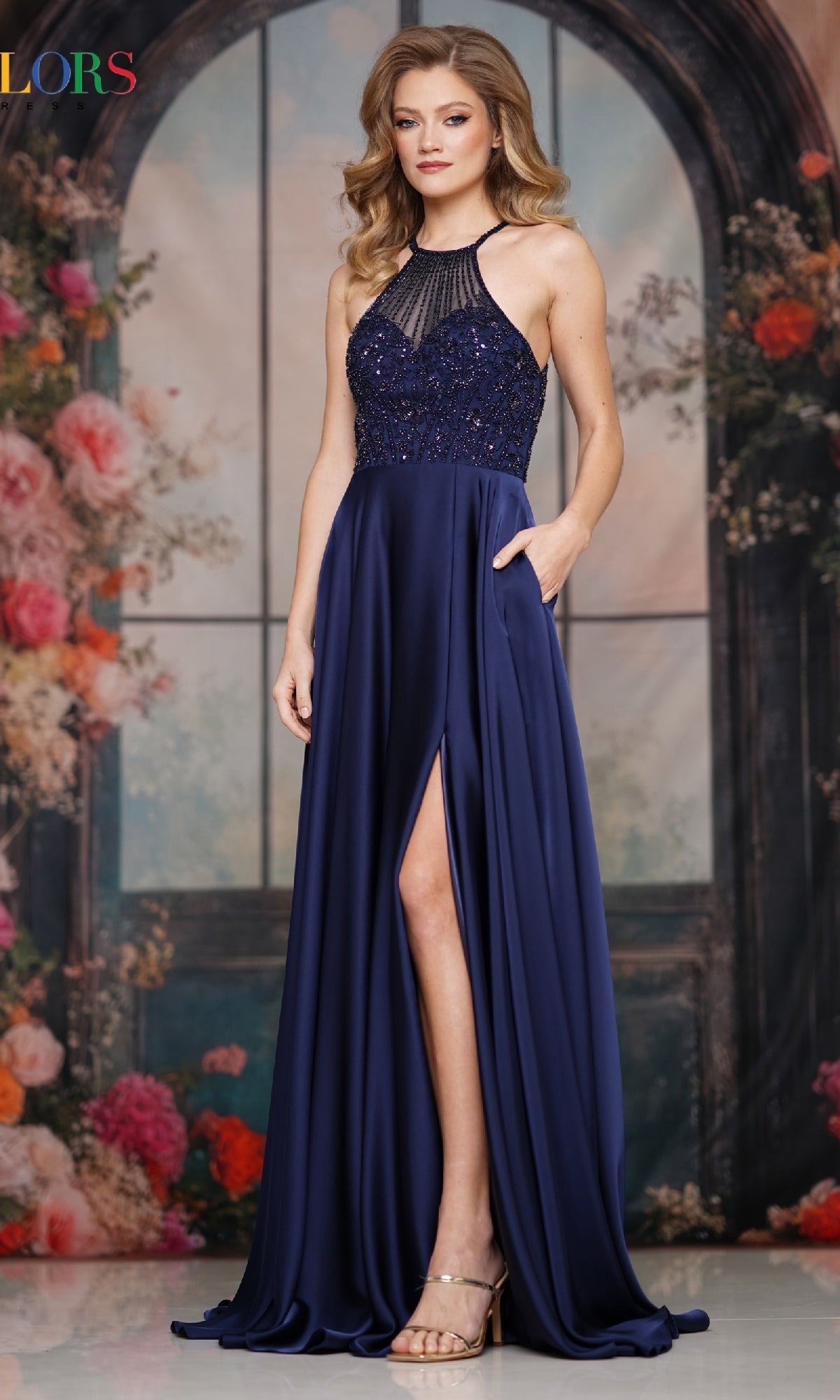 A woman stands before a floral backdrop, wearing gold heels and the Colors Dress 3822—a sleeveless, floor-length navy blue high-neck halter prom gown with a beaded bodice, thigh-high slit, and pockets.